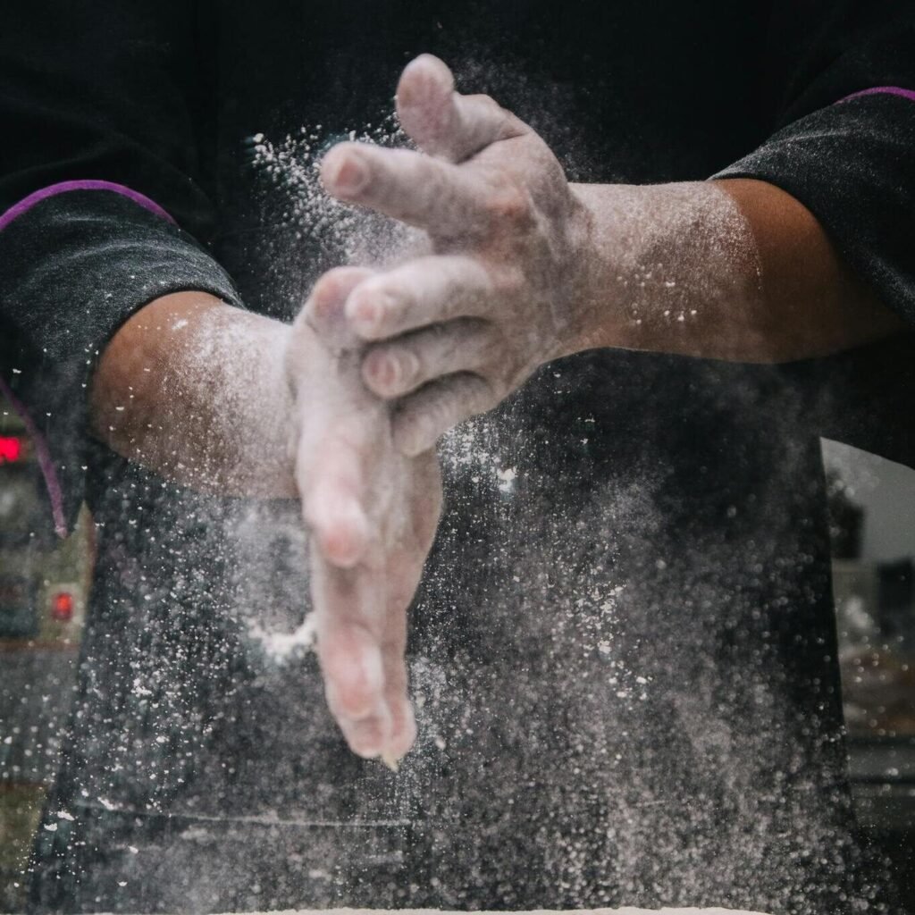 A chef clapping hands over dough, releasing flour in a kitchen setting.